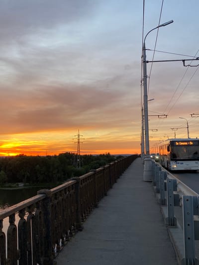 A scenic view of a sunset over Novokuybyshevsk, Russia, captures a picturesque moment on a bridge. The sky is painted with vibrant hues of orange and yellow, casting a warm glow over the landscape. In the foreground, a pedestrian walkway lined with ornate metal railings offers a vantage point to admire the breathtaking sunset. Beyond the walkway, a road features a white bus with illuminated headlights and a destination sign displaying "5п Самарка-Ту". The bus is in motion, heading away from the viewer. On the left side of the image, a river flows gently, reflecting the colorful sky. Lush green trees form a dense forest along the riverbank, adding to the natural beauty of the scene. In the distance, a tall transmission tower stands as a prominent landmark against the twilight sky. The overall ambiance is serene and tranquil, highlighting the beauty of the natural and urban elements coexisting in harmony.