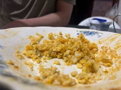 The image shows a partially eaten plate of rice, with a yellow/orange hue, possibly due to spices or sauce. The plate is white with a blue floral design. A small portion of a leg and part of clothing are visible in the lower right corner, suggesting the person is seated. There is a fork and a small package (possibly sauce) on a plate near the bottom left. The setting seems to be indoors. There are no indicators pointing to Ouaouizert, Morocco.