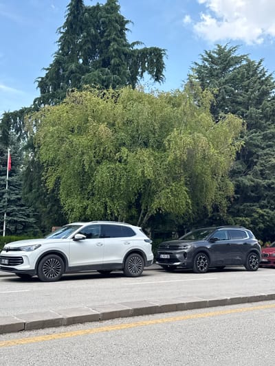 The image shows two cars parked on the side of a road in Etimesgut, Turkey. The car in the foreground on the left is a white SUV. To its right is a dark gray SUV. Further to the right, a portion of a red car is visible. The cars are parked on asphalt with a concrete curb and a yellow line marking the edge of the road. Behind the cars, there are several large trees, including evergreen and deciduous varieties. A red flag on a flagpole is visible to the far left of the image. The sky above is blue with scattered white clouds.