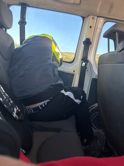 The image depicts the interior of a vehicle, viewed from the rear passenger seats. A person wearing a grey jacket with a high-visibility lime green collar and blue lettering that appears to say "ГРУП-07" is seated in the foreground, facing away from the camera and looking out the window. They are wearing black track pants with white stripes and dark underwear is visible. A black bag with a white splatter pattern is on the seat next to them. The vehicle's interior shows grey seats and a white paneling on the doors. The window reveals a landscape under a clear blue sky, suggesting an outdoor rural setting. The presence of the "ГРУП-07" lettering on the jacket could indicate a team or group designation, and the context of being in a vehicle suggests travel. Without further information, the specific location in Djerman, Bulgaria, cannot be determined from the image alone.