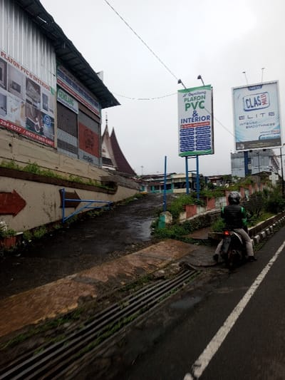 The image depicts an overcast street scene in Chagzam_Pam, Bhutan, characterized by a wet asphalt road and various commercial structures. On the far left, an elevated building with a corrugated metal facade displays signs advertising "PLAFON PVC & INTERIOR" and a business named "GEMERLANG," along with phone numbers. Below this building, a concrete slope leads upward, marked by a painted red arrow and a blue metal barrier. The ground here is uneven and damp, with scattered green vegetation.
In the mid-ground, two large billboards stand prominently. The left billboard, labeled "Gemerlang," advertises "PLAFON PVC & INTERIOR" with blue and green text. The right billboard promotes "NEW CLAS ELITE" with a white and blue design. Distant buildings with distinctive, steeply pitched roofs are visible behind the billboards and to the left of the main slope, suggesting traditional architecture.
On the right side of the main asphalt road, a single individual is seated on a dark motorcycle. The person wears a grey helmet, a dark vest with green shoulder accents, and light-colored pants. The motorcycle is stationary, facing away from the viewer. The road surface is dark and appears wet, featuring a linear drainage grate and a worn white lane marking. Sparse green foliage grows along the road's edge and on low retaining walls. Overhead, a single utility wire traverses the frame against the bright, overcast sky.