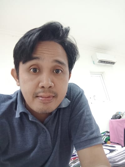 A male individual, appearing to be an adult of Southeast Asian ethnicity, is depicted in a close-up, chest-up shot. He possesses dark hair, styled with volume on top, and exhibits a thin mustache and light stubble on his chin. His eyes are wide, directly engaged with the camera, and his lips are slightly parted. He wears a blue-grey short-sleeved polo shirt with a three-button placket, with one button fastened.

The background reveals an indoor setting with white walls and a white ceiling. A white split-unit air conditioner is mounted high on the wall in the upper right section. Below the air conditioner, a portion of a white-framed window or interior door is visible. In the lower right corner, below the individual's arm, a surface is draped with pink fabric, upon which a grey motorcycle helmet and other indistinct objects are placed. Further down and to the left, a stack of papers or magazines with visible text and graphics is present. The scene is an indoor room, likely an office or personal dwelling. While no specific landmarks or unique architectural features are visible to definitively identify the city as Semarang, Indonesia, the environment is characteristic of a contemporary indoor space in an urban setting.