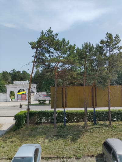 This is a vertical, outdoor, eye-level shot in Iskitim, Russia, showing a bright, sunny day with a clear blue sky. The scene features several tall, slender pine trees with green needles and some bare branches, positioned in front of a yellowish-ochre wall. A neat, green hedge runs along the base of the wall. To the right of the hedge, a patch of rough, sun-drenched grass is visible, with a portion of a light-colored car parked on it.

In the background, an arched white structure, possibly an entrance to a park or garden, is prominent. Above the arch, there's text that appears to read "ГОРСАД ИМ. И.И. ПОРОТЕЕВ" (GORSAD IM. I.I. POROTEYEV). A banner with red and yellow colors is displayed within the arch. Further back, more trees and foliage create a lush, green backdrop. A person is walking away from the camera on a paved path near the white structure.

The image captures a serene and possibly recreational outdoor setting, with the vibrant greenery and clear sky suggesting a pleasant day.