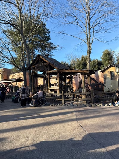 The image displays an outdoor scene within Frontierland at Disneyland Paris in Chessy, France, under a clear blue sky. A prominent rustic, open-sided wooden structure, identified by a facade sign as "BLACKSMITH," stands in the midground. This building houses various metalworking equipment, including large wooden wheels, gears, and machinery, and is partially enclosed by a low wooden fence with a stone base. To the right of the blacksmith shop, a small, light-colored building with a dark roof and an exterior lantern is visible. Additional adobe-style buildings are present in the background.
Approximately eight individuals are depicted. A group of four is situated on the left side of the blacksmith shop: one individual in a plaid jacket and a backpack stands with a walking stick, another is seated in a dark manual wheelchair, and two others are seated nearby. Two indistinct figures are partially visible walking on the far left. A single individual is seated on the ground near the wooden fence on the far right. Mature deciduous trees with bare branches and evergreen pines frame the upper portion of the scene. The foreground consists of a paved asphalt surface with a longitudinal crack. Long, distinct shadows are cast from the left across the pavement, consistent with a low sun angle.