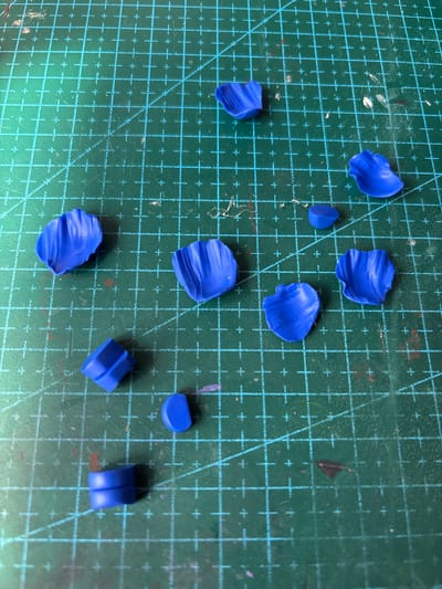 A close-up, overhead shot shows various small, curved, blue objects scattered across a green grid cutting mat. The objects appear to be made of clay or a similar pliable material and are molded into petal-like shapes, some with defined creases and edges. A few of the objects are cylindrical or semi-circular in form. The cutting mat is marked with a grid of turquoise lines, with intersecting horizontal and vertical lines and diagonal lines. Some faint smudges and red marks are visible on the mat. There are no people or animals in the image, and the scene appears to be a tabletop workspace. The image does not provide any information about the location in Blagoveshchensk, Russia.