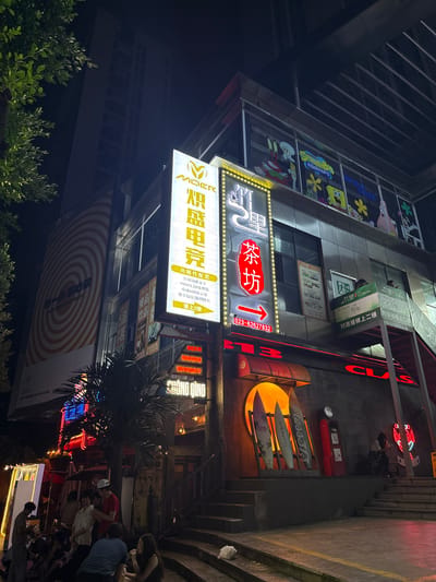 The image depicts a nighttime scene in a bustling urban environment, likely a commercial street in Longta, China. In the foreground, a group of people are gathered outside what appears to be a bar or entertainment venue. They are casually dressed and seem to be socializing. The building behind them is illuminated by a variety of neon signs, indicating various establishments. Notable signs include "BEER," "MUSIC," and "WHISKY." One prominent sign displays "CHONG QING" in large red lettering.
Further up the building, more signage is visible. One large vertical sign features a yellow logo and Chinese characters, with smaller text below detailing "功能性配置" (functional configuration). Another sign with a red background has a white arrow pointing to the right, with Chinese characters that translate to "Tea House" (茶坊). Above this, a sign with a yellow "M" logo appears to be for "MOER" (M.O.E.R.), likely an esports or gaming establishment.
The architecture of the building suggests a modern commercial complex. Windows on higher floors are visible, some displaying colorful graphics, including characters from SpongeBob SquarePants. A large circular advertisement or mural with orange concentric circles is also visible on the side of the building. The overall atmosphere is vibrant and energetic, characteristic of a popular entertainment district at night. Steps lead up to different levels of the building, and palm trees add a tropical touch to the urban landscape.