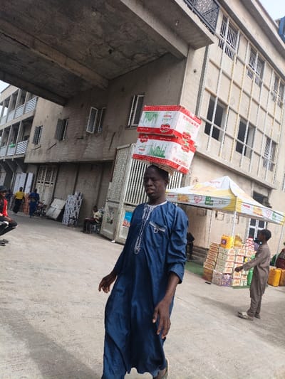 The image depicts an outdoor commercial setting in Fagge C, Nigeria, under daylight. In the foreground, a man of African descent, wearing a dark blue embroidered traditional kaftan, walks forward. He is balancing two stacked rectangular white cardboard boxes, marked with red and green branding including Arabic script and an image of the Taj Mahal, on his head.
The scene features multiple buildings. To the left, a multi-story concrete structure exhibits an exposed concrete overhang and balconies with white railings. Several windows, some open, are visible. Below the overhang, various panels and objects lean against the building. Multiple individuals are present in this area, including one crouching person in a red and black top, another in yellow further back, a person in blue near a motorcycle, and an individual seated under the overhang.
To the right, a multi-story building with a grid of windows and security bars forms the background. A white and yellow canopy tent, branded "Active" with a heart logo, is erected in front of this structure. Under the tent, various packaged goods, including stacks of boxes and yellow containers, are displayed. A young boy in a light brown traditional garment stands near the merchandise, observing it. Yellow plastic jerrycans are visible further to the right. The ground is paved concrete.