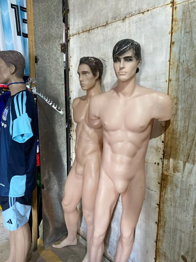 The image shows two nude male mannequins stacked on top of each other, both light-skinned. The top mannequin has dark hair, while the lower one has brown hair. Below them, a clothed mannequin wears a dark blue and light blue Adidas soccer jersey. The mannequins are in a space with metallic and rusted surfaces.