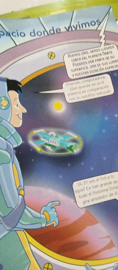 The image depicts an illustration from a children's book or comic, featuring an astronaut in a blue and green spacesuit looking at a celestial scene. The astronaut is on the left side of the frame, facing right, with a smiling expression. They are looking towards a small, light blue spacecraft with yellow accents that is suspended in space. In the background, there is a planet with a reddish-pink hue, a bright yellow sun, and a starry sky.
Two speech bubbles are visible. The first, from the top right, reads: "BUENOS DIAS, HEMOS LLEGADO CERCA DEL PLANETA MARTE. PODEMOS VER PARTE DE SU SUPERFICIE, UNA DE SUS LUNAS Y NUESTRA ESTACIÓN ESPACIAL." This translates to "GOOD MORNING, WE HAVE ARRIVED NEAR THE PLANET MARS. WE CAN SEE PART OF ITS SURFACE, ONE OF ITS MOONS AND OUR SPACE STATION."
The second speech bubble, originating from near the spacecraft, reads: "Gracias, computadora, ¡Qué grande se ve el planeta en comparación con su satélite natural!" This translates to "Thank you, computer, How big the planet looks in comparison to its natural satellite!"
A third speech bubble, located at the bottom right, contains the text: "SÍ, ¿Y VEE el Sol a lo lejos? Es tan grande que todo el Sistema Solar gira alrededor de él." This translates to "YES, AND DO YOU SEE the Sun in the distance? It is so big that the entire Solar System revolves around it."
The overall scene is set in outer space, with a focus on exploration and learning about planets and the solar system. The mood is one of wonder and discovery. The text suggests the characters are exploring Mars. The specific location context of La Tagua, Venezuela, is not visually evident in the illustration itself, but it is provided as external information.