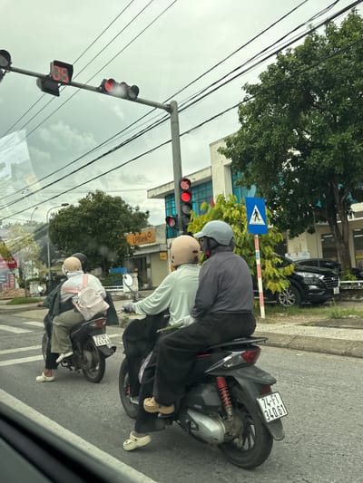 The image depicts a street scene in Triệu Phong Commune, Vietnam. In the foreground, two individuals are riding on a motorcycle, with the passenger in the rear wearing a light-colored backpack. To the left, another motorcycle is partially visible with a passenger wearing a backpack. In the background, a building with an "LPBank" sign is situated, and further back, trees and power lines are visible. A traffic light showing a red signal is positioned overhead, and a pedestrian crossing sign is also present. The sky is overcast.