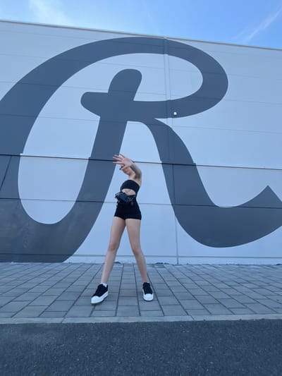 A woman is captured in a dynamic pose against a large, graphic mural on the side of a building. She is wearing a black sports bra, black shorts, and black and white sneakers. Her arms are outstretched and her body is arched backward, showcasing her flexibility. A small black bag with straps is attached to her waist. The mural features a bold, dark grey abstract design with sweeping curves against a white background.
The scene is set outdoors, with a clear blue sky and some wispy clouds visible. The ground is paved with grey bricks, transitioning to a dark asphalt surface. The lighting suggests it is daytime, likely sunny given the bright sky. The overall mood is one of athletic movement and artistic backdrop. The location is identified as Pilsen, Czechia, though no specific landmarks are immediately apparent beyond the building mural and paving. There is no visible text in the image.