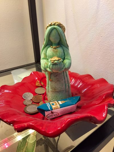 A close-up, eye-level shot shows a figurine of a woman in a green robe with gold accents, holding a gold object in her hands. The figurine stands on a red, petal-shaped dish that contains several coins and a wrapper for candies. The figurine and dish are placed on a reflective surface, possibly a glass table. In the background, a white, textured wall is visible. The scene appears to be indoors, potentially a display or personal collection, with no immediate indication of its location within Arboga, Sweden, or any specific actions or interactions occurring. The lighting suggests an indoor environment.
