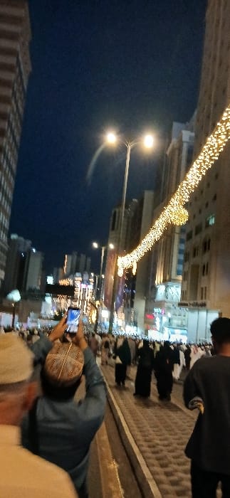 The image depicts a crowded urban street scene at night in Makkah Al Mukarramah, Saudi Arabia. The sky is dark blue, indicating evening. In the immediate foreground, a person wearing a blue garment and a patterned cap is observed from behind, holding a dark smartphone vertically with both hands, aiming it forward. Partially visible to their left is an individual in light-colored attire. To the far right foreground, a person in a dark top walks forward. The street features a textured pavement and a low curb. A dual-lamp streetlight with warm illumination is centered in the mid-ground. Above and to the right, a string of yellow decorative lights hangs diagonally across the street. The background is densely populated with pedestrians, many dressed in traditional Islamic clothing, moving along the thoroughfare. Tall, multi-story buildings, illuminated with windows and architectural lighting, line both sides of the street, extending into the distance.