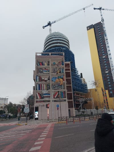 This image captures an urban street scene in daytime under an overcast sky, with several notable architectural and street elements.
**Subjects:** The central focus is a multi-story building on the left adorned with a large, vibrant mural depicting a bookshelf filled with various books. Behind this, a distinctive domed building with horizontal stripes is visible, surrounded by scaffolding with blue tarps, indicating construction or renovation. Further back and to the left, a tall, modern skyscraper with a gold-colored lower section and dark upper floors dominates the skyline. Two large construction cranes are prominently featured in the upper left quadrant, towering over these buildings. On the right, a wide asphalt road includes a red-colored lane with white dashed lines, likely a pedestrian crossing or cycle path, and yellow traffic cones line the curb. Traffic infrastructure includes a red-lit traffic signal, a blue one-way arrow sign pointing left, and a red circular stop sign. A small part of a person's head and shoulder, dressed in dark clothing, is visible in the upper right.
**Activity or event taking place:** The scene depicts typical urban life with ongoing construction and road infrastructure. No specific event is unfolding beyond the daily flow of a city.
**Notable details:** The weather is overcast, resulting in soft, diffused lighting and no harsh shadows. The time of day appears to be daylight. The presence of numerous cranes and scaffolding strongly suggests significant urban development or redevelopment in the area. The "bookshelf" mural is a distinctive artistic feature of the building it covers.
**Visible text:** Text on a sign below the traffic light pole reads "REAL ESTATE" with another word, possibly "CENTER," partially visible beneath it. The titles on the books in the mural are not decipherable.