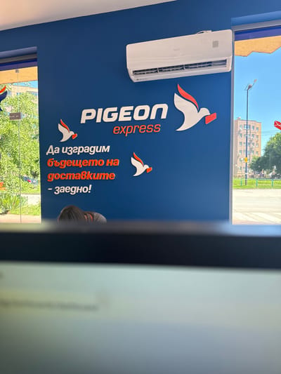 The image shows a storefront with a blue wall featuring the "PIGEON express" logo, which includes a stylized white bird with an orange accent and a red envelope. Below the logo, text in Bulgarian reads "Да изградим бъдещето на доставките - заедно!" which translates to "Let's build the future of delivery - together!". An air conditioning unit is mounted high on the wall. Through a glass door or window to the left, trees and a building are visible, suggesting an outdoor environment. In the foreground, a person's head and shoulders are visible, looking down, as if seated at a desk. The overall scene appears to be an interior view of a business office or service point.

The location of this business in Pazardzhik, Bulgaria can be inferred from the presence of the Bulgarian language in the text on the wall. While the specific street address is not visible, the outdoor scene suggests a commercial area within the city, likely a street-facing establishment. The buildings and street visible through the window are typical of a Bulgarian urban setting.