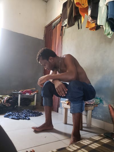 The image, taken with a vertical orientation, captures a young, dark-skinned man indoors. He is shirtless, wearing dark blue shorts, and has short, curly hair. A thin, light-colored chain is visible around his neck. The man is depicted in a bent-over posture, possibly stretching or engaged in a physical activity. His arms are folded across his body, with hands resting near his knees or thighs, and his gaze is directed downwards.
His bare feet are visible on a light-colored, tiled floor. One foot rests on a simple, low wooden stool or frame. The room features light-colored walls, with one section displaying a darker gray hue. To the left of the man, a collection of folded clothes in various colors, including green, yellow, and brown, is stacked on what appears to be a shelf or hanging rack. Behind him, a dark wooden door or door frame is partially visible. On the right side of the frame, a metal or plastic shelving unit holds items, including a red cap. A pile of blue and white striped fabric lies crumpled on the floor near his left foot. Another bare foot, belonging to a different individual, is partially visible in the upper right portion of the image.
The lighting in the room suggests it is daytime, with a bright source of light illuminating sections of the walls and floor. The setting appears to be a casual interior, consistent with a personal living space. No visible text is clearly discernible. The location context for this scene is Atlanta, United States of America.