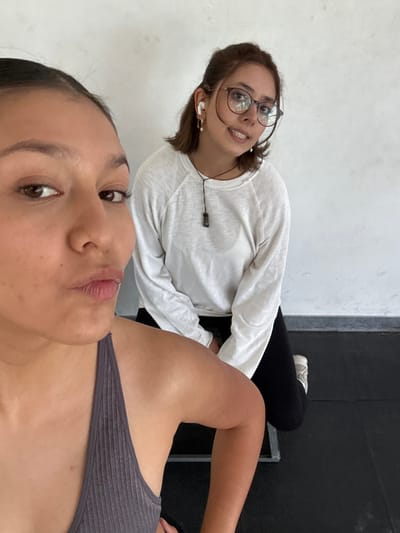 A tilted, close-up selfie captures two young women in an indoor setting, possibly a gym or fitness studio, characterized by a plain white wall and dark flooring or equipment. The image is taken from a low perspective.
In the immediate foreground, partially visible, is the woman taking the selfie. She has dark hair, brown eyes, and full, slightly parted lips. She wears a dark grey ribbed tank top or sports bra, with her shoulder and upper chest visible. Her expression is neutral.
Behind her, looking towards the camera with a relaxed smile, is a second woman. She has brown hair, wears dark-rimmed glasses, and has white wireless earbuds in her ears. She is dressed in a long-sleeved white t-shirt and black pants. A thin black cord is visible around her neck. Her feet, wearing white sneakers, rest on a dark surface, which appears to be a padded bench or mat. The background includes a dark vertical structure, possibly part of gym equipment or a wall trim.
The overall atmosphere is casual and relaxed. No text is visible in the image. The scene is located in San Andrés Cholula, Mexico.