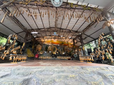 The image depicts a rustic outdoor stage area, likely a performance space or cultural exhibit. The stage is adorned with decorative elements made from natural materials, including dried corn husks and what appear to be bundles of straw or reeds hanging from the roof structure and branches. The background features a faux rock wall adorned with miniature structures, trees, and white goat figurines, suggesting a village or mountainous scene. Several racks display bunches of dried corn. On the left side of the stage, a large red drum is visible. The overall aesthetic is earthy and traditional, possibly representing a rural Vietnamese setting. The "Yang Bay" logo is visible at the top center of the frame, indicating the location is Yang Bay Park in Khanh Vinh, Vietnam. The scene appears to be empty of people.