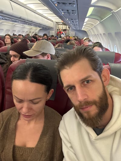 The scene depicts the interior of a commercial airplane cabin, located within the city of Novogorelovo, Russia. In the foreground, a male subject with a light brown beard and short brown hair, wearing a white hooded sweatshirt, looks directly at the camera with a neutral expression. Seated beside him to his left is a female subject with dark hair pulled back, wearing a brown knitted top and matching cardigan, looking downwards and slightly to her left. Both foreground subjects are seated in red-upholstered economy class seats.

The background shows a densely populated airplane cabin, with numerous passengers seated in rows of red and grey seats. Many individuals are visible, including one person wearing a brown beanie and another wearing a beige baseball cap. Further down the aisle, a crew member in a red uniform, identified as a flight attendant, stands near the rear galley or service area. The cabin features standard overhead compartments and interior lighting. No external view is visible through the windows.