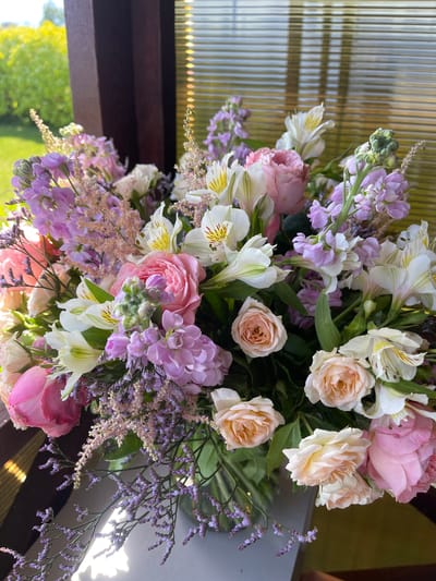 A floral arrangement featuring pink roses, white alstroemeria, and light purple stock flowers is displayed in a vase near a window. The window has blinds, and sunlight is streaming through, creating shadows. In the background, blurred greenery is visible, suggesting an outdoor setting. The image appears to be taken indoors, possibly in a home or event space. The composition focuses on the bouquet, with the window and outdoor scene serving as a backdrop. The photograph is a close-up of the flowers. The location is identified as Zaķumuiža, Latvia.