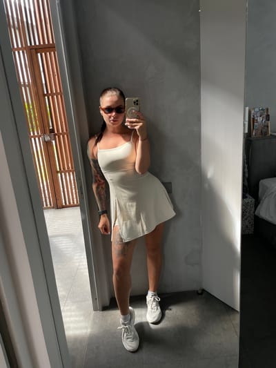 In what appears to be a modern residential interior, possibly a hotel room or private home, a woman is taking a mirror selfie. She stands in a room with gray, textured walls and a white door on the right. To her left, a wooden slatted door or screen is visible, casting striped shadows on the floor. The woman is wearing a cream-colored, spaghetti-strap dress with a thigh-high slit, white athletic sneakers with white socks, and tortoiseshell sunglasses. Her hair is pulled back in a braid, and she has extensive tattoos on her left arm and leg. She is holding a smartphone in her right hand, capturing her reflection. The lighting suggests it is daytime, with sunlight entering the room. The environment is clean and minimalist, with a bed and nightstand visible in the background to the right. There is no direct indication of the location being in Kediri, Indonesia, but the architectural style and general ambiance could be consistent with contemporary designs found in various urban settings, including potentially in Indonesia.