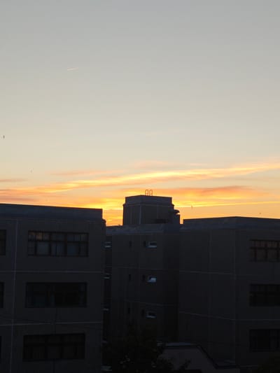 The image depicts several buildings in the foreground silhouetted against a vibrant sunset. The buildings are of a utilitarian design, possibly residential or institutional. The windows are dark, suggesting the interior lights are off or the windows are not reflecting much light. The sky exhibits bands of orange, yellow, and pink hues, indicating a sunset or sunrise. The upper portion of the image is a lighter shade, transitioning into the sunset colors. A contrail is visible in the upper left. The location is likely Markovskii, Russia, based on the prompt. There is no indication of human activity or interaction in the scene.