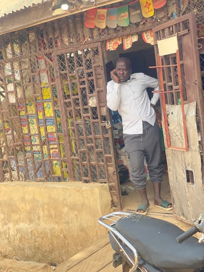 A male individual stands in the doorway of a small commercial establishment in Unguwar Gabas, Nigeria. He is wearing a light-colored, vertically striped long-sleeved shirt, dark grey cargo-style sweatpants, and light green slide sandals. He holds a dark mobile phone to his right ear with his right hand, with his left hand resting on his hip. He is looking forward, slightly to the right of the viewer.
The shop entrance features a rusted brown metal security gate or grille, partially open. To the right of the entrance is a metallic door or panel. A padlock is visible on the security gate. Above the entrance, a corrugated metal roof is visible, along with an exposed light bulb. Below the roofline, a horizontal string displays various empty product sachets, including brands such as "MILO," "NESCAFE," and "MAGGI."
Through the open gate, the shop interior reveals stacks of yellow and blue packages, possibly powdered milk, and several large white sacks. Blue plastic containers are also visible within the shop.
The exterior ground directly in front of the shop is unpaved earth. A raised concrete platform forms the base of the shop entrance. In the lower right foreground, the rear section of a dark-colored motorcycle or scooter is visible, including its black seat with a silver grab rail.