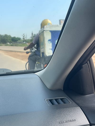 A person on a bicycle with a large cooler box on the back is visible from inside a car. The person is wearing a dark shirt and grey pants, and a yellow helmet. The cooler box is white with blue accents and appears to have a logo or branding on it. The bicycle is dark colored. The scene is outdoors, with a paved road and some vegetation visible in the background. The sky is clear and blue. The interior of the car, including the dashboard and a vent, is in the foreground. The image appears to have been taken in Kuchingoro, Nigeria.