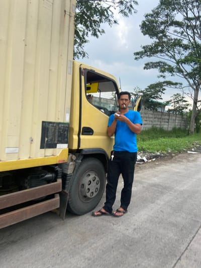 A man in a blue shirt and black pants stands next to a yellow truck. He is holding a cigarette in his right hand and appears to be smoking it. The truck is parked on the side of a paved road. The road is surrounded by vegetation. The man is looking directly at the camera. The location is Margaasih, Indonesia, based on the provided coordinates (latitude: -6.969, longitude: 107.549). The image appears to have been taken during the day, as the sky is clear and the sun is shining. The truck's cab door is open and a hand can be seen inside, possibly reaching for something. There are keys attached to the truck's side. The road is made of concrete, and it is dusty. The pavement is slightly cracked and uneven. There is a small amount of litter on the side of the road, near the truck. There is a concrete wall and a row of trees in the background.