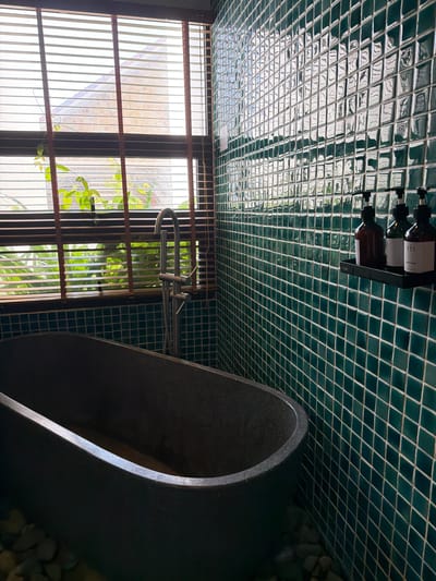 The image depicts a modern bathroom featuring a deep, dark gray bathtub situated next to a window covered by horizontal wooden blinds. The bathtub appears to be made of a textured material, possibly stone or a similar composite. Green, square mosaic tiles cover the wall behind the bathtub, creating a vibrant and reflective surface. On a small black shelf mounted on the tiled wall are three dark-colored pump bottles, likely containing toiletries. Outside the window, lush green foliage is visible, suggesting a connection to nature or an outdoor garden. The overall aesthetic is one of contemporary luxury with a touch of natural elements. The scene is set in a private bathroom within South Quy Nhon Ward, Vietnam.