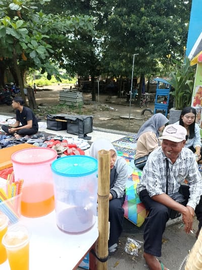 This image captures a vibrant outdoor market or gathering in Mataram, Indonesia, under the shade of numerous large trees. The scene is bustling with people and various stalls.
In the foreground, a makeshift stall offers refreshments, featuring two large plastic drink dispensers – one pink filled with an orange liquid, and another blue one containing a purple beverage. Several cups of a yellow drink, likely juice, and a container of colorful straws are also visible. A bamboo pole supports this stall.
Several individuals are seated on the ground, which is partially covered by a colorful patterned mat. On the far right, an older man with a warm smile, wearing a white cap and a checkered shirt, sits comfortably. Beside him, a woman in a grey hijab is looking towards the right. Further back, another woman in a beige hijab and a woman with dark hair in a light blue top are also seated. To the far left, a young man in a black t-shirt and pants sits cross-legged near an array of sandals and other footwear laid out for sale, suggesting a footwear stall. A black duffel bag is placed on the ground behind him.
The background reveals a dirt ground area with more trees, suggesting a wider open space. Several motorbikes are parked, and indistinct figures can be seen in the distance, further indicating an active public space. A blue pushcart or small stall is visible under the trees on the right, though its contents or any text on it are illegible. The bright daylight suggests it is either morning or afternoon, and the overall atmosphere appears relaxed and social. An empty cigarette pack and a cigarette butt are seen on the ground near the older man.
