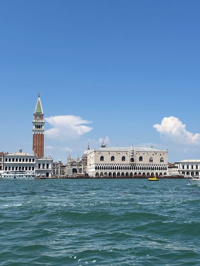 The image depicts a panoramic view of the Venetian lagoon with a historic cityscape lining the water's edge. The buildings are predominantly white and ornate, featuring arched windows and intricate architectural details, suggestive of Venetian Gothic style. Prominently featured is a tall, slender bell tower with a square base and a conical, green-tiled roof, topped with a spire. Adjacent to it, a grand, multi-story building with a long colonnade runs along the waterfront. Another imposing structure, likely the Doge's Palace, with its distinctive facade, is also visible. The water is a deep turquoise, rippling with small waves, and a bright yellow object, possibly a buoy or a small boat, is floating near the buildings. The sky is a clear, vibrant blue with a few scattered, fluffy white clouds. The sunlight suggests it is daytime, likely late morning or early afternoon, creating bright highlights on the buildings and the water. The scene conveys a sense of tranquility and historical grandeur, characteristic of Venice. There is no visible text in the image.