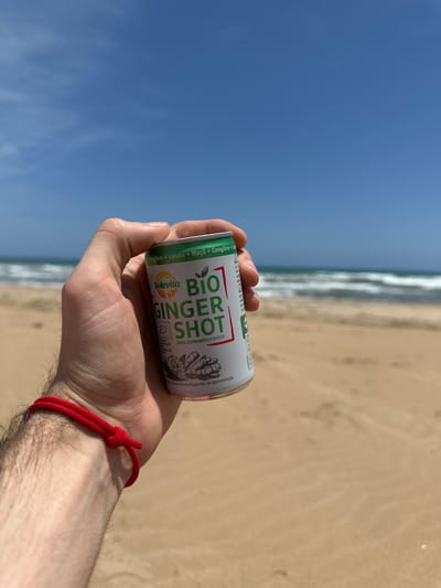 A close-up, first-person perspective photograph captures a fair-skinned individual's right hand holding a small aluminum can on a sunny beach in Guardamar del Segura, Spain. The hand, exhibiting moderate hair on the forearm, wears a red string bracelet tied with a simple knot on the wrist. The can, predominantly white with green and red accents, displays the brand "Solevita" and the product name "BIO GINGER SHOT." Visible text indicates "Gengibre + Limón + Maçã + Gengibre" (Ginger + Lemon + Apple + Ginger) and "100% EXPRIMIDO/FRESCO" (100% SQUEEZED/FRESH), along with an organic certification logo. The background features a sandy beach with golden-brown sand, extending towards the ocean. Light blue-green water with breaking white waves is visible in the middle distance. The horizon is flat, separating the ocean from a clear, vibrant blue sky that dominates the upper portion of the frame, showing only a few faint, wispy clouds in the far upper left.