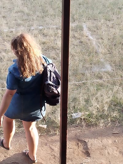 A woman with curly, light brown hair pulled back in a ponytail wears a denim dress and a dark backpack while standing outside. She is seen from the back, wearing sandals and stepping towards the left. Behind her, a grassy field stretches out, with a white painted line visible in the distance. A weathered, dark brown pole bisects the frame, partially obscuring the view. A discarded plastic bottle rests on the ground near the fence.