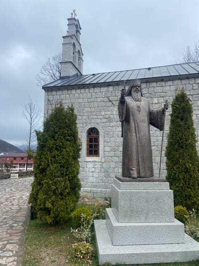 The image shows a bronze statue of a robed figure, possibly a religious figure, standing in front of a stone building. The statue is positioned on a tiered granite base. Behind the statue is a stone building with a tower and an arched window. Two tall, green, coniferous trees flank the statue. A stone-paved path leads away from the statue, and a building with red and white elements is visible in the background. The sky is overcast. This location is in Kolašin, Montenegro.