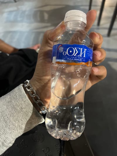 A person is holding a nearly empty, clear plastic water bottle with a white cap. The bottle has a blue and orange label with the brand name "ИЗО" and other text in Hebrew and Arabic. The person's hand and wrist are visible, with a silver chain bracelet on their wrist. The person is wearing a light grey long-sleeved shirt. The background is out of focus, showing hints of a patterned black surface and a person in a black shirt. Given that "BAS" is written on the bottle, it is possible this image was taken in Skhirat, Morocco as the company of the same name has headquarters there.