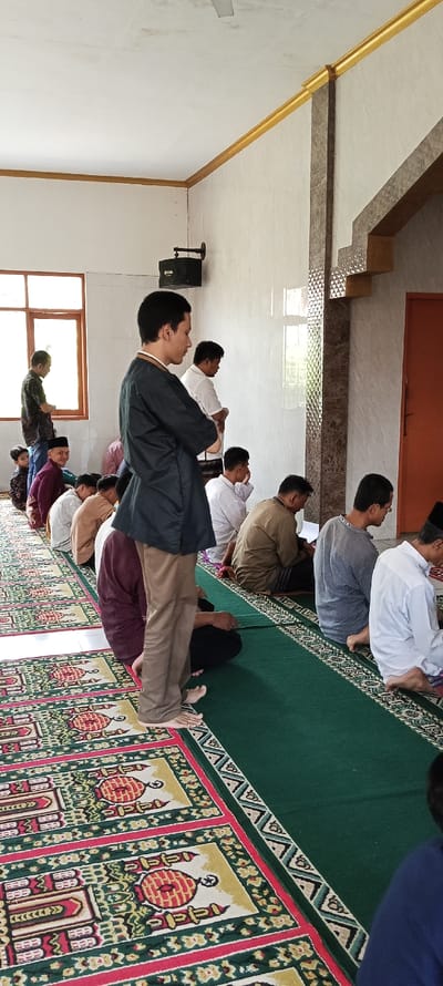 The image shows a room with a green carpet covering the floor. There are several men sitting on the carpet, with one man standing in front of them. The men are all wearing long-sleeved shirts and pants. The man standing in front of the group is wearing a black long-sleeved shirt and brown pants. The carpet is covered with prayer rugs, with intricate designs and patterns of red, yellow, and green. The walls of the room are white and decorated with a brown border.  The room is well-lit and the walls are clean. 

The location of the image is a room in a mosque in Kecamatan Kutawaringin, Indonesia, based on the provided coordinates (-6.973, 107.521).