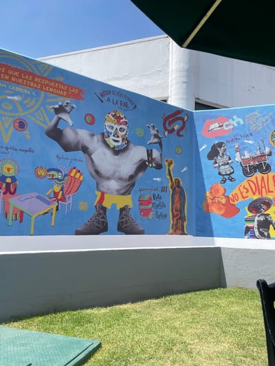 This image features a vibrant, multi-panel mural adorning an exterior wall in San Andrés Cholula, Mexico, under a clear, bright blue sky. The artwork, executed in a colorful, collage-like style, celebrates Mexican culture and linguistic diversity.
The upper mural panel includes text like "NO ES DIALE" (likely "NO ES DIALECTO"), alongside images of marigolds, a traditional figure, and a vintage camera. Phrases such as "llévele, llévele" (a vendor's call) and references like "PACHUCOS RIFAN" (Pachucos rule) and "CHENDO" suggest cultural pride and local expressions.
The larger lower panel is dominated by a grayscale luchador (Mexican wrestler) with a colorful mask, arms outstretched. To his left, bold text reads, "NADIE LE PIDE PERMISO A LA RAE. DEJEN COTORREAR" (No one asks permission from the RAE. Let them chat), a defiant statement about linguistic freedom from the Royal Spanish Academy. This section also includes "OS QUE LAS RESPUESTAS LAS EN NUESTRAS LENGUAS" (We have the answers in our languages) and "In quihtoa moyollo," likely an indigenous language phrase. Whimsical figures like a red serpent and various colorful creatures are scattered throughout.
To the luchador's right, a depiction of the Virgin Mary holding flowers appears above a section titled "sparinglish 101," providing examples like "Rufa (TECHO)," "Marketa (SUPERMERCADO)," and "Parkear (ESTACIONAR)," humorously illustrating Spanglish. A "SUPERMENSA" box is also visible. Other text like "Aguitado, comadre" (down/sad, friend) adds to the mural's playful and culturally rich narrative.
The scene is brightly lit by strong midday sun, casting defined shadows. To the right of the mural, a strip of artificial grass is visible, suggesting an outdoor patio or public space. A green umbrella can be seen partially on the far left. The mural champions the richness of local languages, slang, and cultural identifiers, reflecting a strong sense of identity.