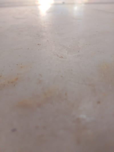 The image shows a close-up, out-of-focus view of a light-colored, textured surface. The surface appears to be flat, possibly a tabletop or floor, with subtle variations in color and some small, dark specks scattered across it. The lighting suggests a bright environment, with highlights reflecting off the surface, creating a blurred, elongated glow at the top of the image. There are no identifiable objects, people, or discernible environmental features that would indicate a specific location within Peshawar City Tehsil, Pakistan. The overall impression is of an abstract texture rather than a recognizable scene.