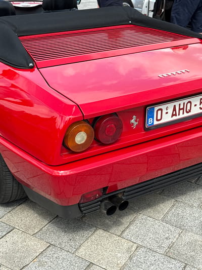 This is a close-up, low-angle, medium shot of the rear of a red Ferrari, possibly a classic model. The car is parked on a cobblestone street. The Ferrari's red paint gleams, reflecting the sky. Prominently displayed is a Belgian license plate that reads "0 - AHQ - 5" with a blue European Union circle and a "B" for Belgium. A small silver prancing horse emblem, the Ferrari logo, is visible just below the license plate.
To the left of the license plate, a black convertible top is partially visible, suggesting the car is a convertible. The rear of the car features two distinct taillights: an upper circular red taillight and a lower circular amber light. To the right of the taillights, two chrome exhaust pipes are visible, protruding from the rear bumper.
The background shows a textured cobblestone ground, characteristic of historic European cities like Bruges. The lighting suggests it's daytime, possibly overcast, as there are soft reflections on the car's body. No people are directly visible, but the presence of the car and its parking location imply it's part of a street scene, possibly during a car show, a tourist attraction, or simply a parked vehicle in a city. The overall impression is one of automotive elegance and a classic European setting.