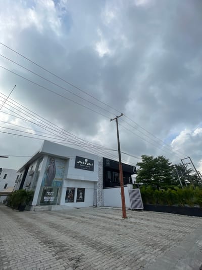 The image shows a building with the sign "Make Me! Beauty Place" in Moba, Nigeria. There is an advertisement for "SWELLS BRAD Duchess" and another sign with the text "We Define it MIZANI" in the window. The sky is cloudy and there are power lines visible in the foreground. The street in front of the building is paved with stone blocks. Some trees and greenery are visible next to the building.