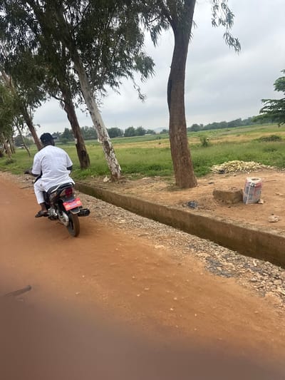 In the city of Katambe, Nigeria, a man wearing a white kaftan and a black cap rides a black motorcycle down a reddish-brown dirt road. He is positioned in the center left of the frame, facing away from the camera. The motorcycle has a red license plate with white lettering and a prominent exhaust pipe on the right side. Beside the road, a concrete drainage channel runs along the right side of the frame. Beyond the channel, an area of dry, yellowed grass is visible, with a pile of cut sugarcane stalks and a bag of cement sitting on the dirt ground. Further in the background, a field of green crops stretches towards a line of trees under an overcast sky. Several tall, slender trees with visible bark patterns line the left side of the dirt road, their leaves creating a dappled shade. Two figures are distantly visible in the green field.