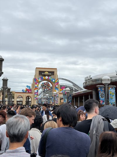 The image shows a large crowd of people gathered outdoors under an overcast sky, likely at the entrance to an amusement park. The perspective is from within the crowd, looking towards a prominent arched gateway in the midground.

The subjects are predominantly the backs and sides of numerous individuals, dressed in casual clothing. Their hair colors vary, with some showing dark hair, others lighter, and a few with graying hair. Faces are generally not visible, except for one woman looking back towards the camera from the left side of the frame.

The central feature of the scene is a beige-colored arched entrance structure, clearly marked with the words "UNIVERSAL STUDIOS." Below this text, the arch is adorned with colorful, stylized cloud and balloon-like decorations and a large "25" insignia, indicating a 25th-anniversary celebration. Beyond this arch, parts of buildings and what appears to be a roller coaster track are visible.

To the right of the arch, several vertical banners are displayed, featuring cartoon characters (including Minions) and repeating the "UNIVERSAL STUDIOS" and "25" motifs. The weather is cloudy, suggesting a mild, possibly cool, daytime. The overall impression is one of a busy, anticipated event at Universal Studios, consistent with its location in Osaka, Japan.