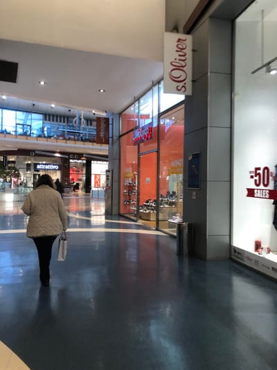 The image captures an interior view of a modern shopping mall in Marousi, Greece. A person, viewed from behind, is walking away from the camera along a wide concourse. The individual is wearing a light-colored, possibly quilted, jacket and dark trousers, carrying a white shopping bag in their right hand.
The mall features a dark, reflective floor with white dividing lines and light-colored walls and ceilings. On either side of the concourse, there are various retail stores with large glass display windows. The store on the left, partially visible, has a sign reading "s. Oliver" in red script. Further down on the left, another store sign reads "Moo&co" (upside down).
On the right side of the concourse, a store window displays merchandise like shoes and clothing. A large banner within this display, seen upside down, advertises "50% SALES". Below this, another store sign for "attraettivo" is clearly visible, and another partially visible sign says "lymac". Reflections of the overhead lighting and store interiors are visible on the polished floor and glass surfaces. A silver trash bin is also visible near the stores on the right.
The scene depicts a typical shopping environment, with artificial lighting illuminating the area. The activity is that of a person navigating the mall, likely engaged in shopping.