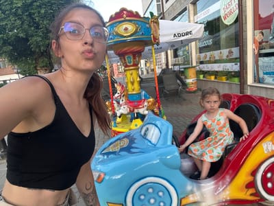A young girl with blonde hair, wearing an orange and white dress with an orange fruit pattern, is seated in a red and yellow toy car. She is looking towards the camera. Beside her, a blue toy car with cartoonish eyes and a flames design is visible, part of a carousel. In the foreground, a woman with brown hair, wearing a black tank top and glasses, is taking a selfie. Her lips are pursed, and she is looking towards the camera. The carousel has small white horses with black spots. In the background, a building with shop windows displaying food and promotional posters is visible. One poster shows cakes and fruits, another advertises "DELICIOUS" and "FAST FOOD". A banner reads "ADDRE". Another sign says "FAST WORLD FAST FOOD". The scene appears to be outdoors during the daytime, under good weather conditions, likely in a public area like an amusement park or a street with shops. The overall mood seems playful and happy, centered around a carousel ride.