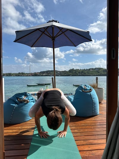 A person is practicing yoga in a crane pose on a wooden deck overlooking a body of water. The deck has a wooden plank floor and is furnished with two large blue beanbag chairs. A blue umbrella is open and positioned behind the person. The water is calm and appears to be in a bay or lagoon, with a distant shoreline featuring greenery and buildings. Two small boats are visible on the water. The sky is partly cloudy, with blue sky visible. The scene suggests a tropical or subtropical location. The specific location is in Nusa Penida, Indonesia.