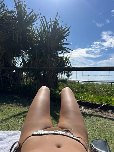 This image, originally rotated 90 degrees counter-clockwise, depicts a bright outdoor scene with a focus on leisure by the coast. In the foreground, the bare, tanned legs and lower torso of a person are visible, suggesting they are relaxing or sunbathing. They are wearing a bikini bottom adorned with iridescent, disc-shaped embellishments. A white towel is partially visible beneath them on the right.
The person is positioned on a patch of vibrant green grass, which extends to a dark wire mesh fence with wooden posts. Beyond this fence, there is a strip of dense, wild green vegetation, characteristic of coastal flora, including what appears to be pandanus palms with their distinctive spiky leaves. Through and above this vegetation, a light blue body of water, likely the ocean, is visible in the distance, indicating a beachfront setting.
The sky in the background is a clear, bright blue with scattered white, wispy clouds. Strong sunlight illuminates the scene, creating highlights on the person's skin and casting shadows on the grass. The overall impression is one of a serene, sunny day at a coastal location, such as Coolangatta, Australia. No discernible text is visible in the image.