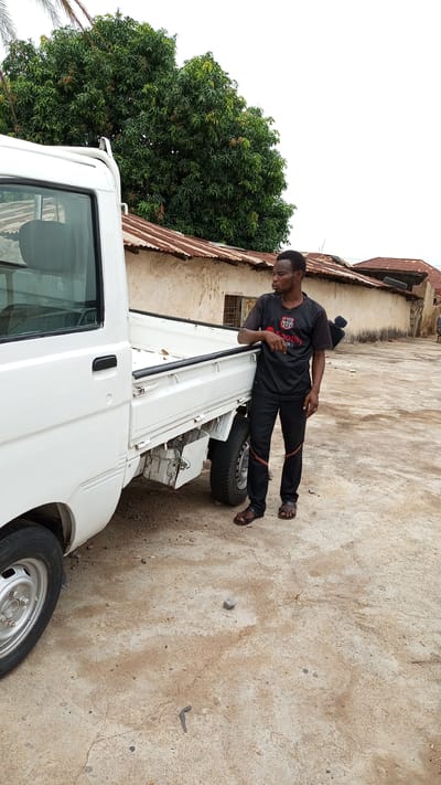 The image shows a man standing next to a white pickup truck in Kawo, Nigeria. The man is wearing a black jersey, black pants with orange stripes, and brown sandals. He is leaning against the side of the truck, looking to his left. The truck has a metal frame and exposed rear tires. The ground is concrete and there are small rocks scattered around. In the background, there is a building with a rusty corrugated metal roof and a tree with dense green foliage.