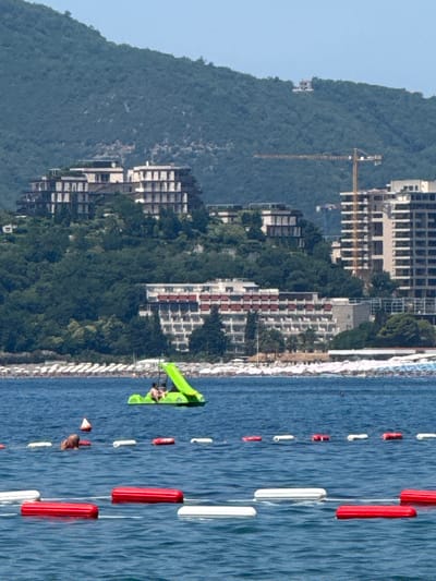 The image depicts a scenic coastal view in Kamenovo, Montenegro, characterized by a lush green mountainside overlooking a beach and the azure Adriatic Sea. Several unfinished or under-construction buildings, likely hotels or residential complexes, are visible on the slope, with a prominent construction crane suggesting ongoing development.
The foreground of the image is dominated by the sea, marked by a series of red and white buoys forming a safety barrier or channel. A bright green inflatable slide is partially visible on the water, with a few people enjoying it. One person is clearly seen on the slide, and another is partially submerged in the water.
The beach itself is lined with rows of white sun loungers, indicating a popular tourist destination. The clear blue sky suggests fair weather, likely during daylight hours, perhaps mid-morning or afternoon. The overall impression is one of a vibrant coastal resort area undergoing expansion, with visitors enjoying recreational activities by the sea. No visible text is present in the image.