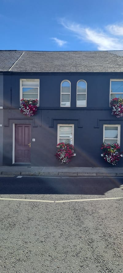 The image shows the facade of a dark blue building with several windows and a dark red door. The building appears to be an apartment or commercial building, as indicated by the multiple windows stacked vertically. Some of the windows have flower boxes with colorful flowers. The sky above is blue with some wispy white clouds. To the right of the building is an asphalt road with a white line marking its edge. A shadow from an unseen object falls across the road. The scene appears to be set on a sunny day. There is a small sign with the number "74" above the door. The overall impression is a residential or commercial street in a town or city. The image is oriented vertically, so the building appears to be on the left and the road on the right.